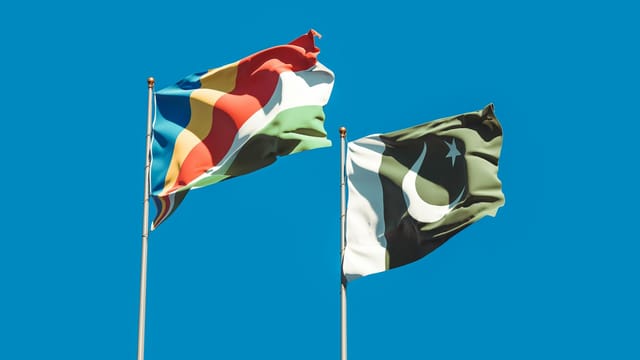 National flags of Pakistan and Seychelles waving on tall flagpoles against a clear blue sky.