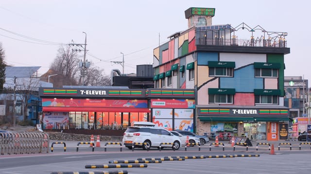 A vibrant 7-Eleven store with colorful facade and parking lot in the foreground, during daytime.