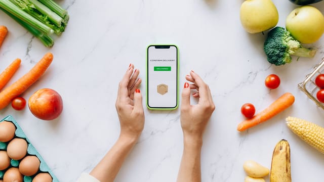 Hands holding smartphone displaying online grocery order amidst fresh vegetables and fruits.