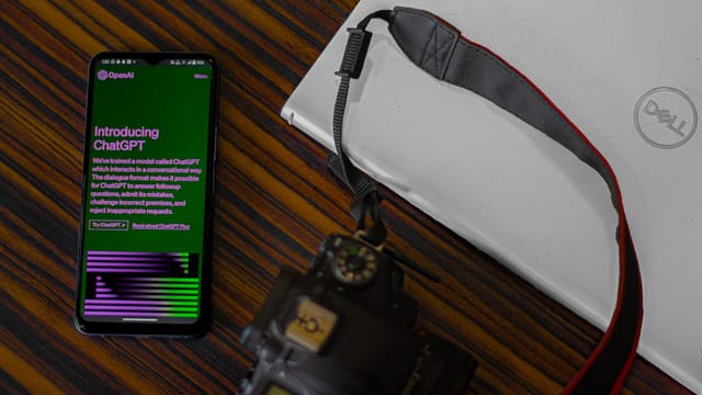 Smartphone with ChatGPT screen next to camera and laptop on wooden desk.
