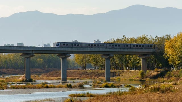 A modern train traverses a bridge with a scenic mountain backdrop near Beijing, China.