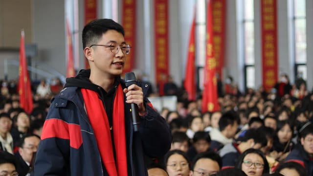 A young man speaking at a crowded indoor event, symbolizing public engagement.