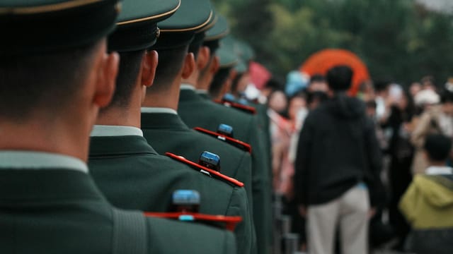 Military officers in green uniforms stand in line formation outdoors, facing a crowd.