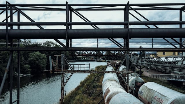 An industrial scene with railway tank wagons and overhead pipes by a canal.