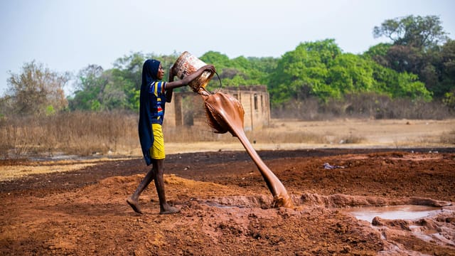 A young girl pouring muddy water from a bucket on a field in rural Nigeria, highlighting local life.