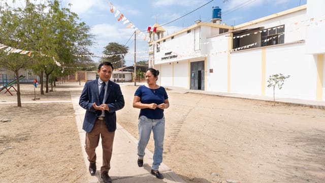 Two adults walk along a pathway outside a building in sunny Piura, Peru, on a clear day.