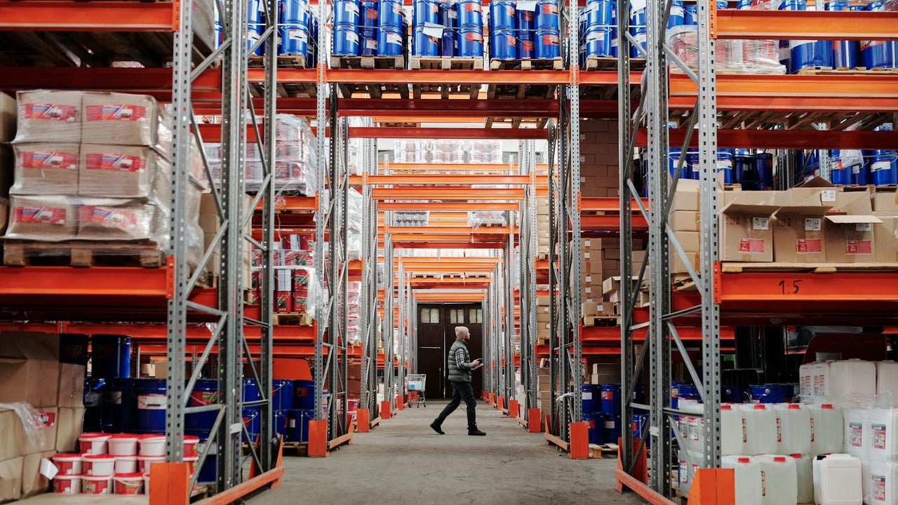 A man walking through a large industrial warehouse with stacked shelves filled with goods and products.
