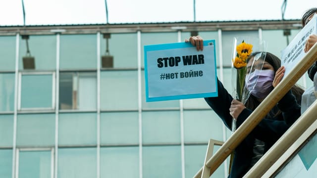 A woman with a mask holds a 'Stop War' sign and a sunflower at an outdoor protest.