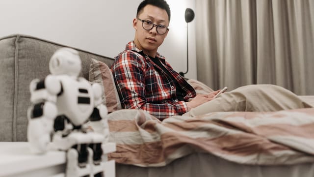 Asian man in plaid shirt sitting on bed, observing a robot toy in a cozy bedroom setting.