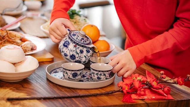 Crop unrecognizable female pouring hot beverage from ornamental teapot into cup at table with oriental dishes in house