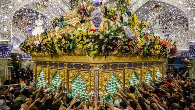 Crowded scene of devotees reaching for a beautifully adorned Islamic shrine under shimmering lights.