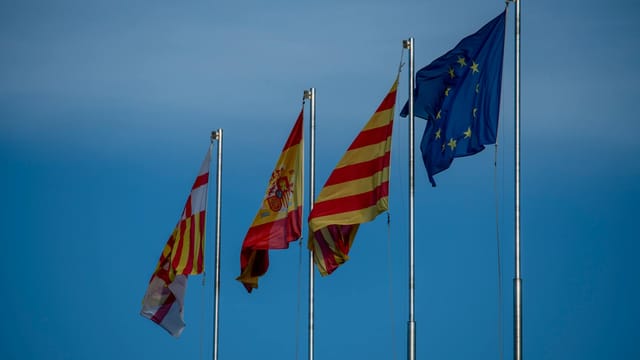Flags of Spain, Catalonia, and Europe on flagpoles against a blue sky in Barcelona.