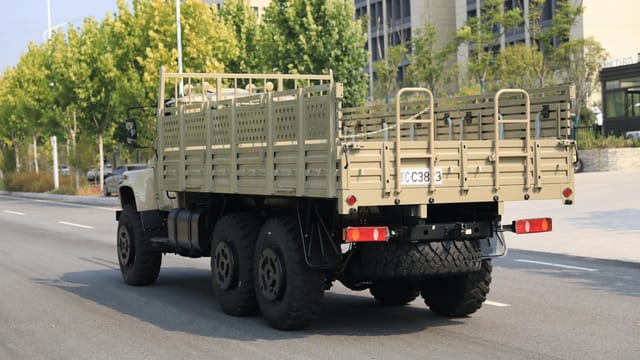 A rugged military cargo truck driving on an urban street lined with trees and buildings.