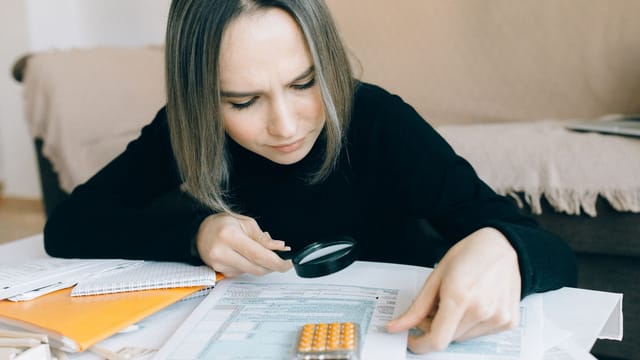 A focused young woman examines documents with a magnifying glass, highlighting attention to detail in accounting tasks.