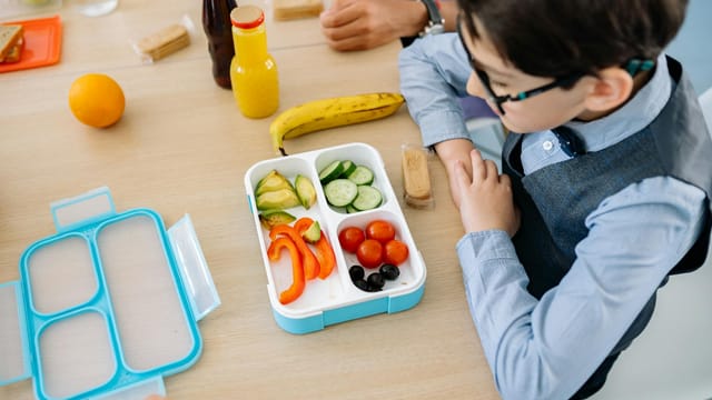 A young boy sits at a wooden table in school, enjoying a healthy lunch from a colorful lunchbox.