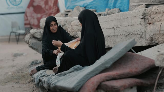 Two women in hijabs sit on rubble in Gaza, sharing a moment of resilience.