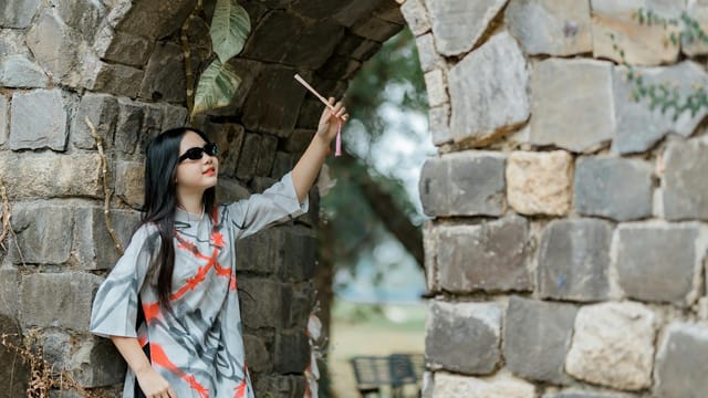 Woman in stylish dress and sunglasses reaches out near a stone archway in an outdoor setting.
