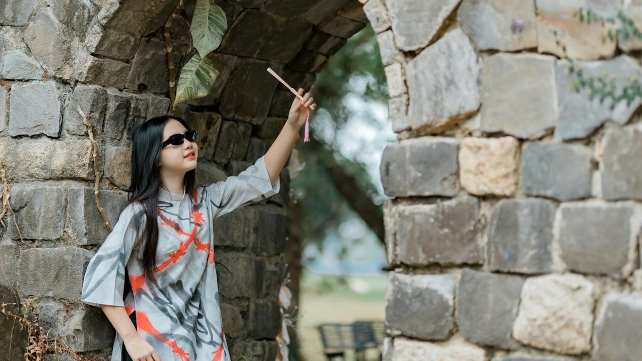 Woman in stylish dress and sunglasses reaches out near a stone archway in an outdoor setting.