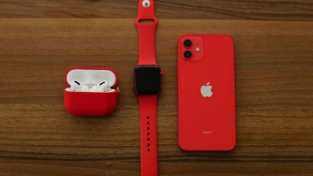 Red Apple iPhone, AirPods, and Apple Watch arranged on a wooden table.