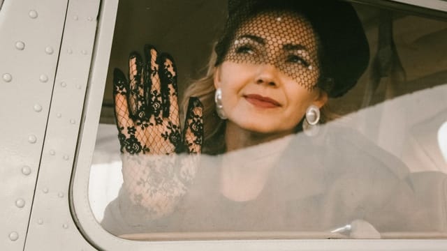 A stylish woman in lace gloves and hat peers through an airplane window with a serene expression.