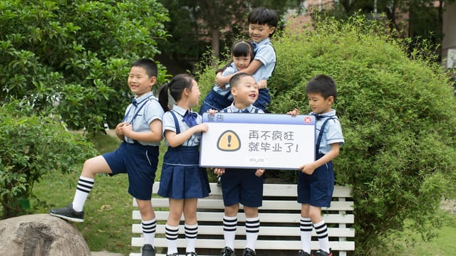 Children in school uniforms having fun outdoors, holding a playful sign on a bench.