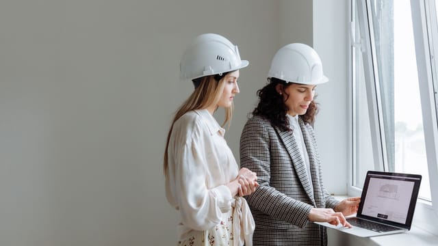 Two female professionals in hard hats review construction plans on a laptop by a window.