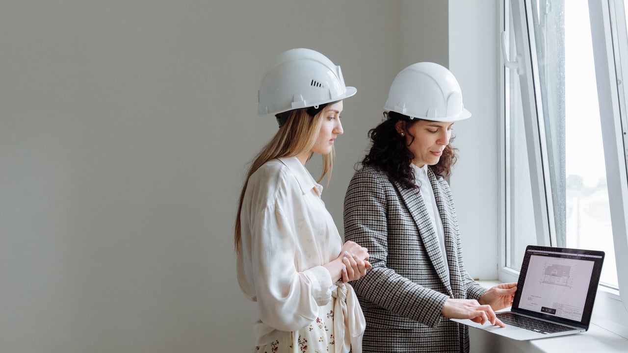 Two female professionals in hard hats review construction plans on a laptop by a window.