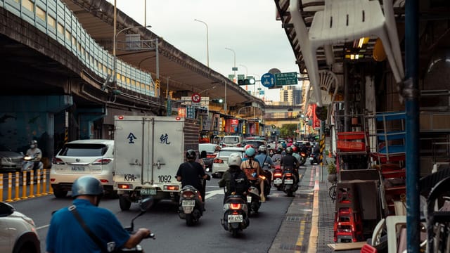 A bustling city street filled with motor scooters and vehicles under an overpass, capturing urban life.