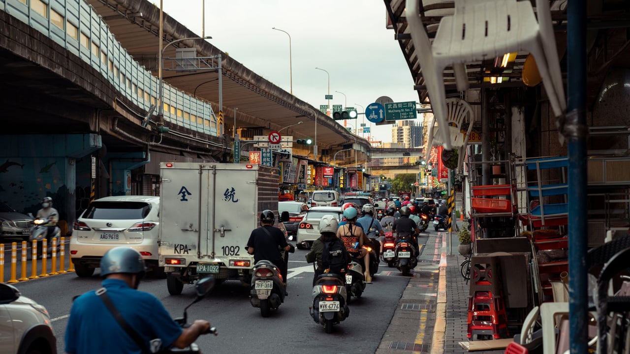 A bustling city street filled with motor scooters and vehicles under an overpass, capturing urban life.