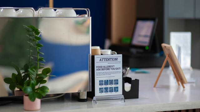 Coffee shop counter featuring an allergy warning sign and plant, highlighting food safety.