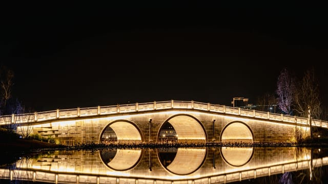 A beautifully lit arched bridge reflecting on a calm river at night in Chengdu, China.
