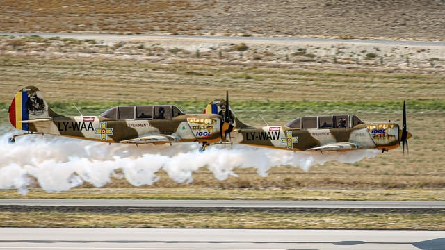 Two Yakovlev Yak-52 planes in synchronized flight with smoke trails over a field.