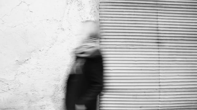 Blurred motion of a person walking past a textured urban wall in monochrome.