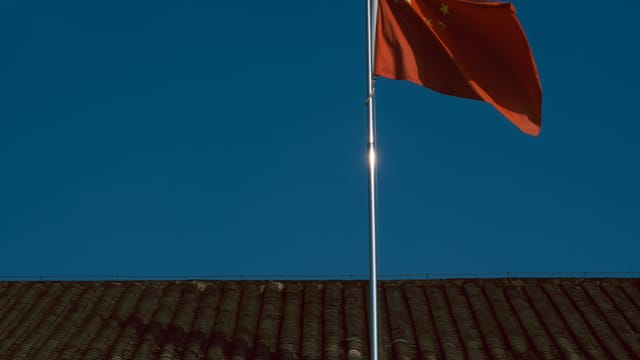 Chinese flag waving over traditional building roof against a clear blue sky.