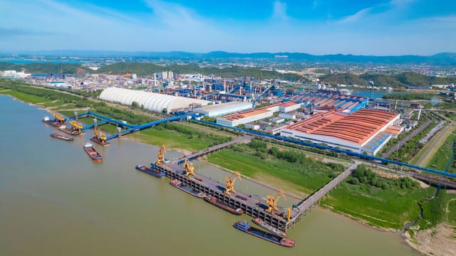 A spacious industrial port with yellow cranes, warehouses, and river access under a clear blue sky.