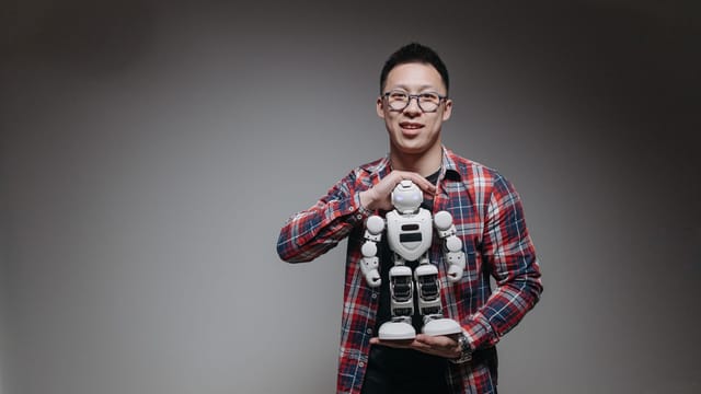 Asian man with eyeglasses holding a toy robot in a studio with a gray background.