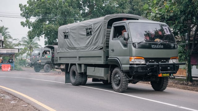 Military trucks traveling on a rural road surrounded by trees and local buildings.