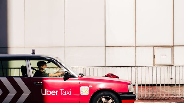A red Uber taxi drives on a sunlit street in Hong Kong's urban landscape.