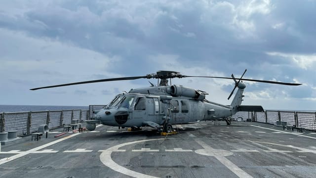 A military helicopter on a naval flight deck under a cloudy sky at sea.