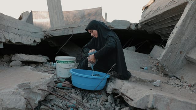 A woman in traditional attire washes clothes among the rubble in Gaza, depicting resilience.