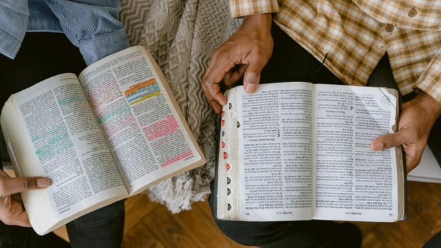 Two adults reading religious texts with highlighted passages indoors, sharing a moment of learning and connection.
