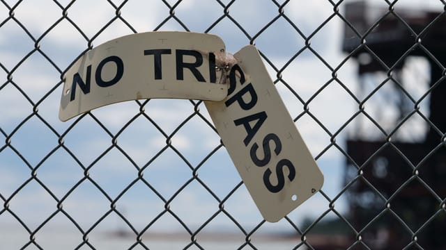 Close-up of a broken 'No Trespassing' sign on a chain-link fence by the water.