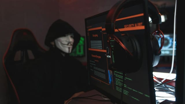 A person wearing a Guy Fawkes mask engaged in hacking activities on a computer in a dimly lit room.