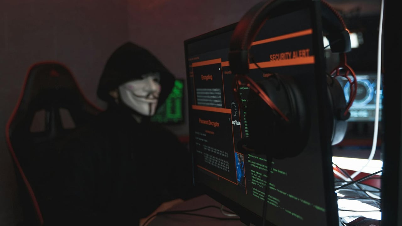 A person wearing a Guy Fawkes mask engaged in hacking activities on a computer in a dimly lit room.