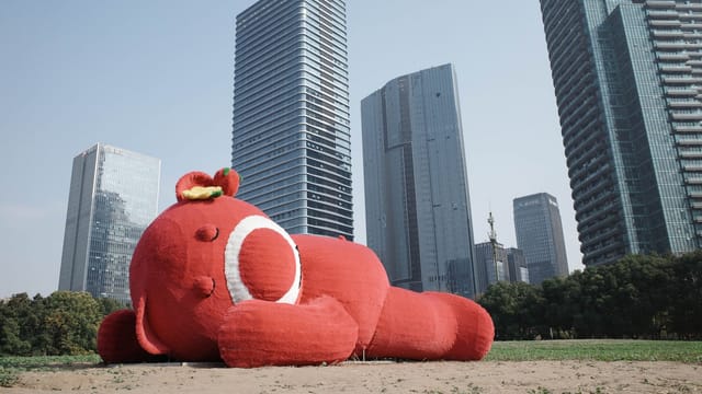 A large red toy sculpture lying on grass with towering skyscrapers in the background under a clear sky.