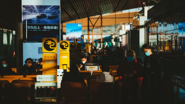 Crowded airport terminal scene with passengers wearing face masks and signage in multiple languages.