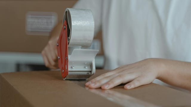 A person sealing a cardboard box with tape, emphasizing packaging and shipping processes.