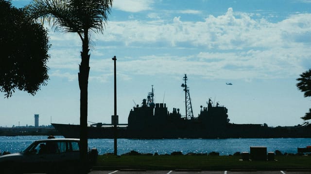 Silhouette of a navy warship in daylight, framed by palm trees and a clear sky.