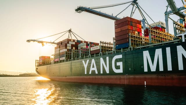 Large cargo ship with stacked containers and cranes at sunset in a harbor.