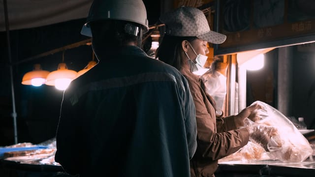 Two people in safety gear interact at a vibrant night market food stall.